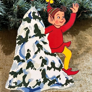 May include: A vintage Christmas decoration depicting a cartoon elf in a red outfit, yellow legs, and red shoes, waving. The elf is positioned beside a snowy tree, topped with a blue and green bird. The ornament is set against a backdrop of evergreen branches.