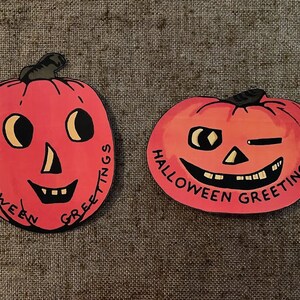 Puede incluir: Dos recortes de papel de calabaza roja con ojos, narices y bocas negras. El texto "Halloween Greetings" está escrito alrededor del borde de cada recorte.