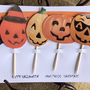 May include: A set of 7 Halloween cupcake toppers featuring orange and black jack-o'-lantern faces with different expressions. The toppers are on wooden sticks and have the text "Happy Halloween" and "Non-Toxic Handmade" printed on the bottom.