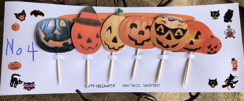 May include: A set of 7 Halloween cupcake toppers featuring orange and black jack-o'-lantern faces with different expressions. The toppers are on wooden sticks and have the text "Happy Halloween" and "Non-Toxic Handmade" printed on the bottom.