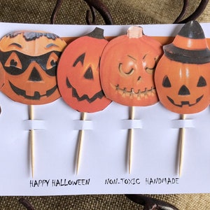 May include: Set of six Halloween cupcake toppers featuring various pumpkin faces with different expressions. The toppers are made of paper and have wooden sticks attached to the back. The card reads "Happy Halloween" and "Non-toxic Handmade".