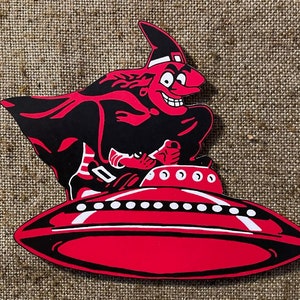 Puede incluir: Una bruja de dibujos animados roja y negra con un sombrero puntiagudo vuela en un platillo volante rojo con detalles blancos.