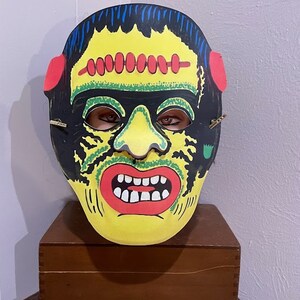 May include: A vintage Halloween mask of Frankenstein's monster. The mask is made of cardboard and features a yellow face with green and black details, red lips, and white teeth. The mask has a black hairpiece and red ears.