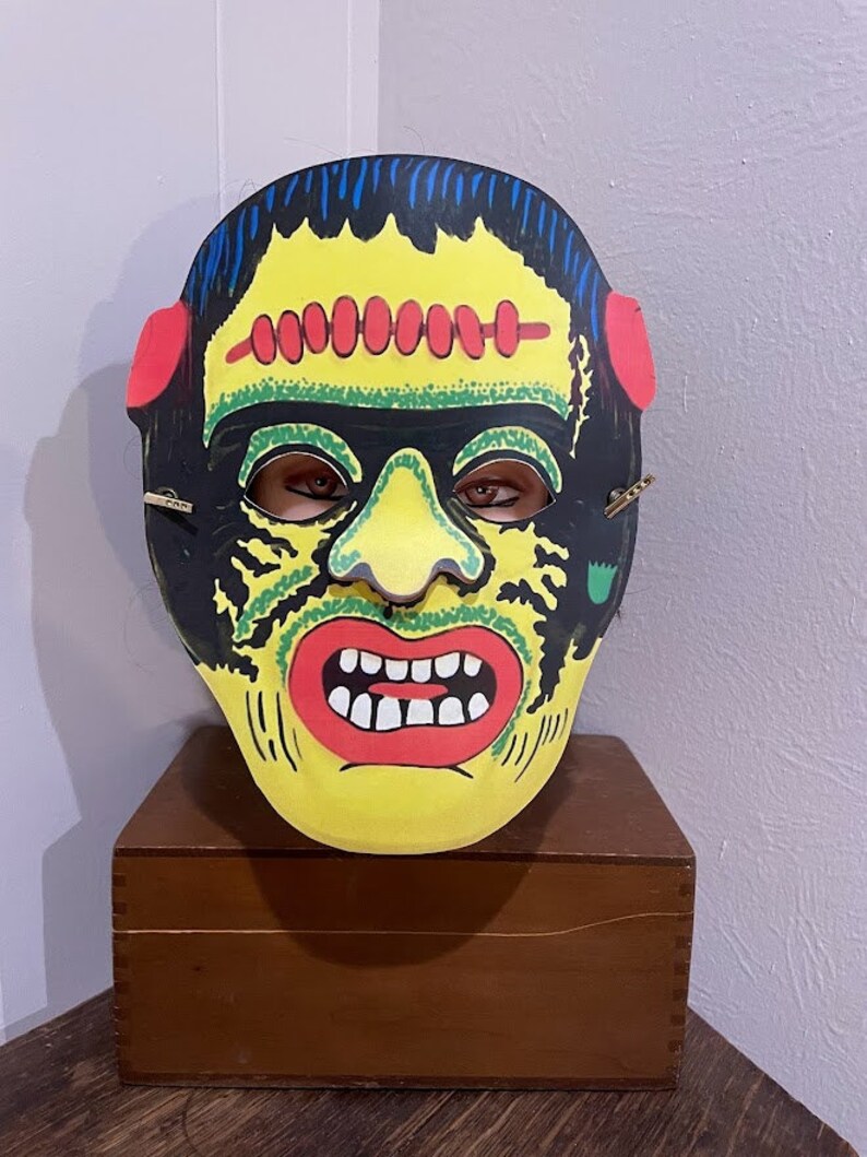 May include: A vintage Halloween mask of Frankenstein's monster. The mask is made of cardboard and features a yellow face with green and black details, red lips, and white teeth. The mask has a black hairpiece and red ears.