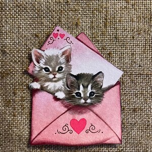以下が含まれることがあります： ピンクの封筒に、2匹の子猫が顔を出しています。封筒には赤いハートと装飾的な渦巻き模様があります。