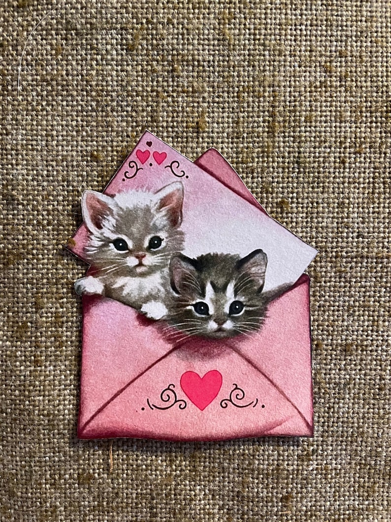 以下が含まれることがあります： ピンクの封筒に、2匹の子猫が顔を出しています。封筒には赤いハートと装飾的な渦巻き模様があります。