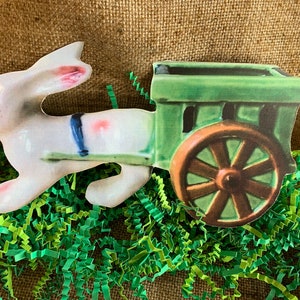 May include: A white porcelain bunny pulling a green wagon with a brown wooden wheel. The bunny has pink ears and blue accents. The wagon has a green wooden body and a brown wooden wheel.