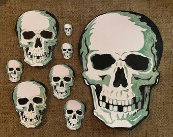 Könnte beinhalten: Ein Set mit acht weißen und grünen Totenkopf-Aufklebern. Der größte Totenkopf hat einen Durchmesser von etwa 15 cm und eine schwarze Umrandung. Die kleineren Totenköpfe haben einen Durchmesser von etwa 2,5 cm.