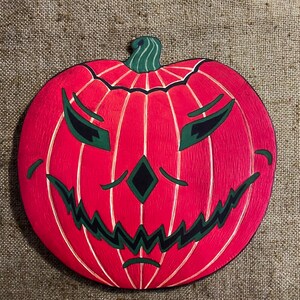 Puede incluir: Una calabaza de madera tallada con una cara de miedo. La calabaza es roja y verde, con una amplia sonrisa de dientes y grandes ojos oscuros.