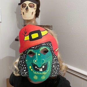May include: A child wearing a green and black witch mask with a red and yellow hat. The mask has a wide, toothy grin and green warts.