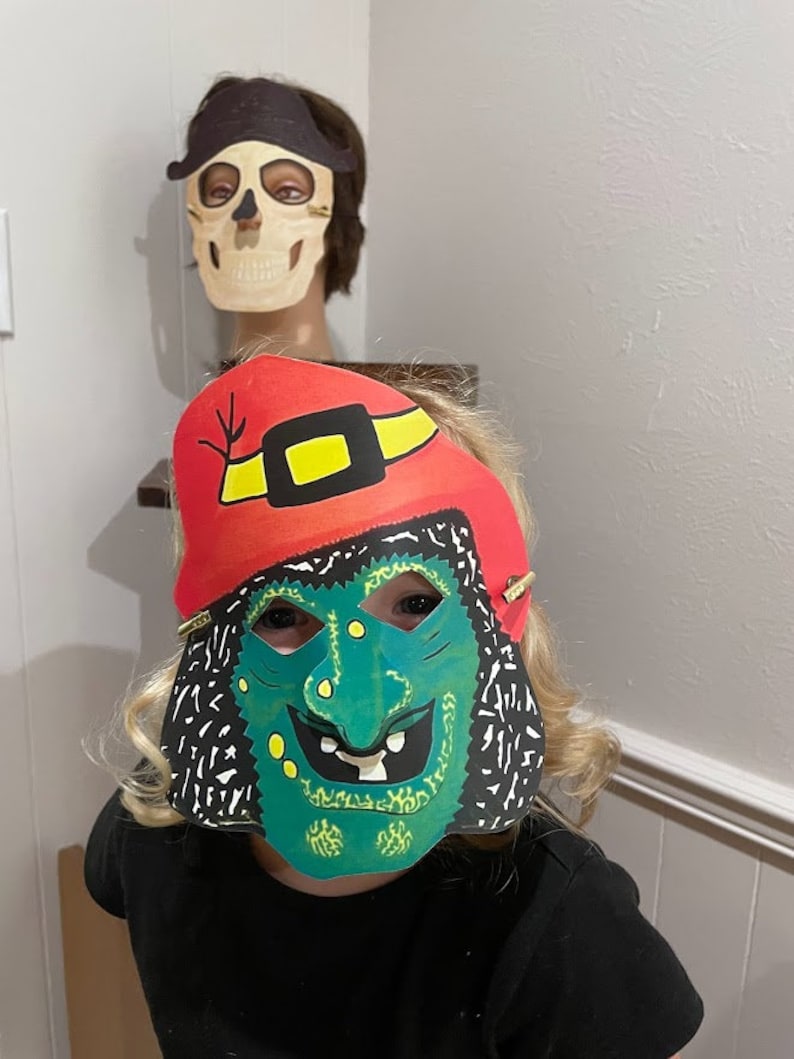 May include: A child wearing a green and black witch mask with a red and yellow hat. The mask has a wide, toothy grin and green warts.