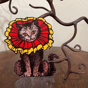 May include: A black cat wearing a red and yellow ruffled collar. The cat is sitting on a wooden surface.