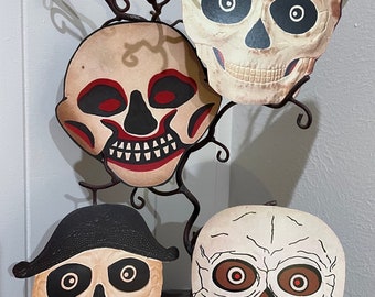 Könnte beinhalten: Drei vintage Halloween-Schädelmasken mit schwarzen Augen und roten und weißen Details. Die Masken sind aus Papier und hängen an einem schwarzen Metallzweig.