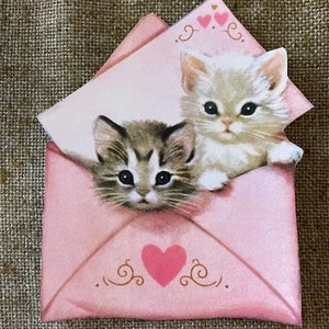 以下が含まれることがあります： 2匹の子猫が顔を出したピンクの封筒。封筒にはハートと装飾的な渦巻き模様があります。封筒のテキストには「愛」と書いてあります。