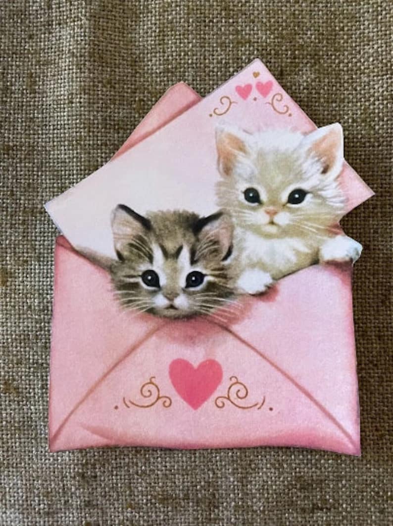 以下が含まれることがあります： 2匹の子猫が顔を出したピンクの封筒。封筒にはハートと装飾的な渦巻き模様があります。封筒のテキストには「愛」と書いてあります。