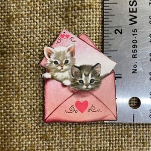 以下が含まれることがあります： 2匹の子猫が顔をのぞかせているピンクの封筒。封筒には赤いハートと「愛してる」の文字が書かれている。