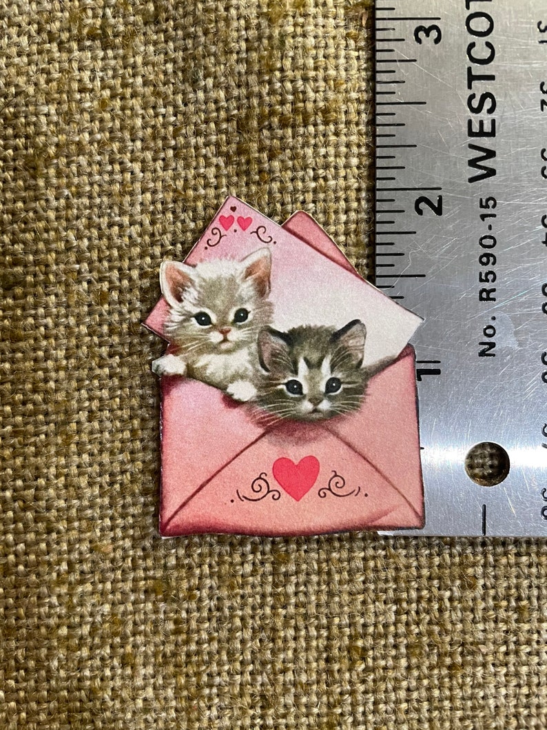 以下が含まれることがあります： 2匹の子猫が顔をのぞかせているピンクの封筒。封筒には赤いハートと「愛してる」の文字が書かれている。