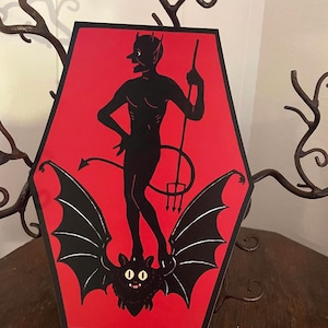 May include: A red and black illustration of a devil standing on a bat with a trident. The image is in the shape of a coffin.