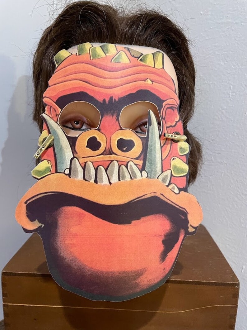 May include: A red and yellow monster mask with large teeth and tusks. The mask has a detailed design with a textured surface.
