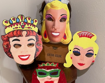 May include: Four colorful paper masquerade masks with crowns and eye makeup. The masks are yellow, red, and white with black eyelashes and red lips. The masks are on a wooden table with a brown box that says "Extra Value".