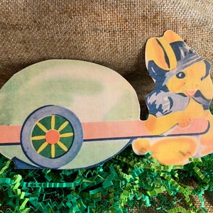 May include: A vintage paper Easter craft depicting a yellow bunny pulling a green and blue wagon. The bunny is wearing a blue hat and has a pink nose. The wagon has a single large wheel with a yellow and green design.