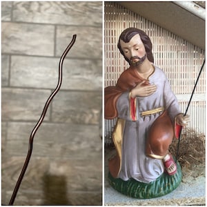May include: A figurine of a man in a brown and white robe, holding a staff. He is kneeling with his head bowed, as if in prayer. The figurine is set against a backdrop of a wooden structure with bamboo reeds.