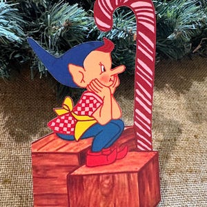 Puede incluir: Un adorno navideño con un duende de dibujos animados con orejas y pantalones azules, una camisa a cuadros rojos y blancos y zapatos rojos. El duende está sentado en un bloque de madera junto a un bastón de caramelo a rayas rojas y blancas.