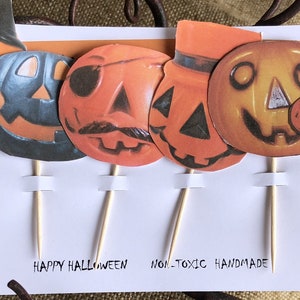 May include: Set of six Halloween cupcake toppers featuring various pumpkin designs with different facial expressions. The toppers are made of paper and have wooden sticks. The text "Happy Halloween - Non-Toxic Handmade" is written on the white background.