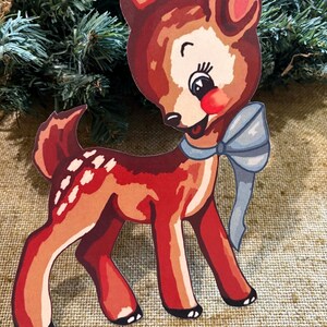 Puede incluir: Un recorte de ciervo de estilo vintage con un lazo azul claro. El ciervo es principalmente rojo y marrón con manchas blancas en la espalda. Tiene una nariz roja y una expresión alegre. El fondo incluye ramas de hoja perenne y una superficie similar a la arpillera.