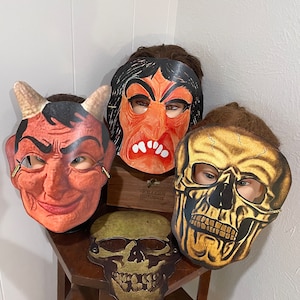 Puede incluir: Cuatro máscaras de Halloween vintage, incluyendo un diablo, una bruja y dos calaveras. Las máscaras están hechas de papel y tienen un acabado pintado. Las máscaras se exhiben en una mesa de madera.