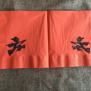 May include: Two orange paper napkins with black silhouettes of witches flying on broomsticks. The napkins have a scalloped edge.