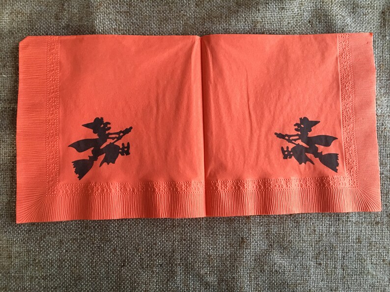 May include: Two orange paper napkins with black silhouettes of witches flying on broomsticks. The napkins have a scalloped edge.