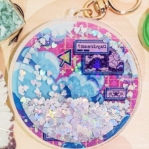 May include: A clear, round keychain with a gold clasp. The interior features a pink grid background with blue clouds, a computer screen that says "Daydreams", and iridescent heart-shaped glitter. The keychain is filled with small, silver heart-shaped confetti.