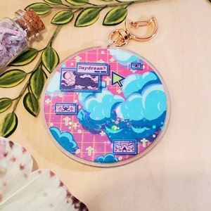 May include: A round keychain with a dreamy design. The keychain features a pink grid background with blue clouds, stars, and computer-like windows. The text "Daydream?" is visible. A gold-colored clasp is attached.