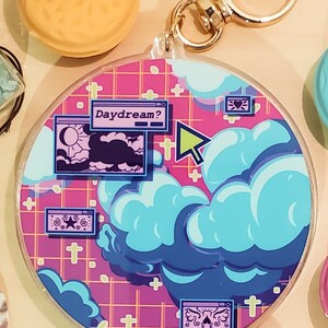 May include: A round keychain with a pink grid background and blue and purple clouds. The keychain has a gold clasp and features the text "Daydream?" in a computer window design. A green cursor points towards the design.