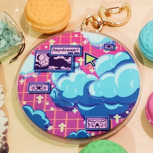May include: A round, clear acrylic keychain with a colorful design featuring blue clouds, a pink grid background, and the word "Daydream?". The keychain has a gold-colored clasp and is surrounded by decorative macarons and crystals.