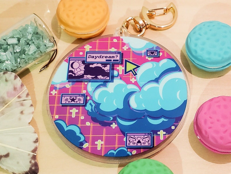 May include: A round, clear acrylic keychain with a colorful design featuring blue clouds, a pink grid background, and the word "Daydream?". The keychain has a gold-colored clasp and is surrounded by decorative macarons and crystals.