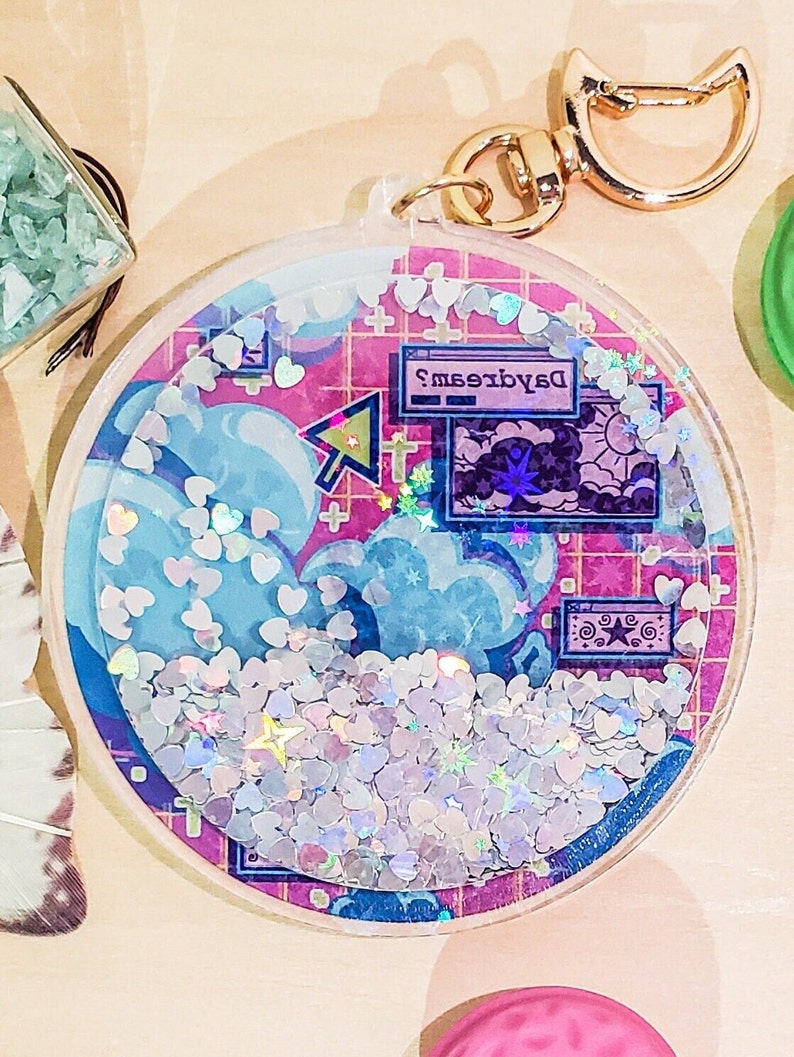 May include: A clear, round keychain with a gold clasp. The interior features a pink grid background with blue clouds, a computer screen that says "Daydreams", and iridescent heart-shaped glitter. The keychain is filled with small, silver heart-shaped confetti.