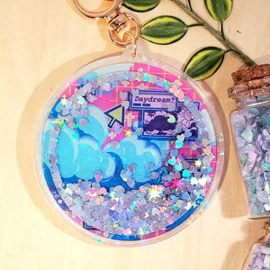 May include: A round, clear acrylic keychain with a gold-colored clasp. The keychain features a blue and pink design with clouds, a computer cursor, and the text "Daydream?". The interior is filled with iridescent glitter.