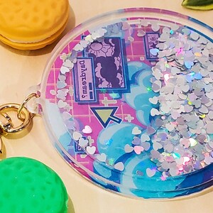 May include: A clear, round keychain with a gold clasp. The keychain features a design with blue clouds, pink grid, and heart-shaped glitter. The text "Dysgessw" is visible on the design.