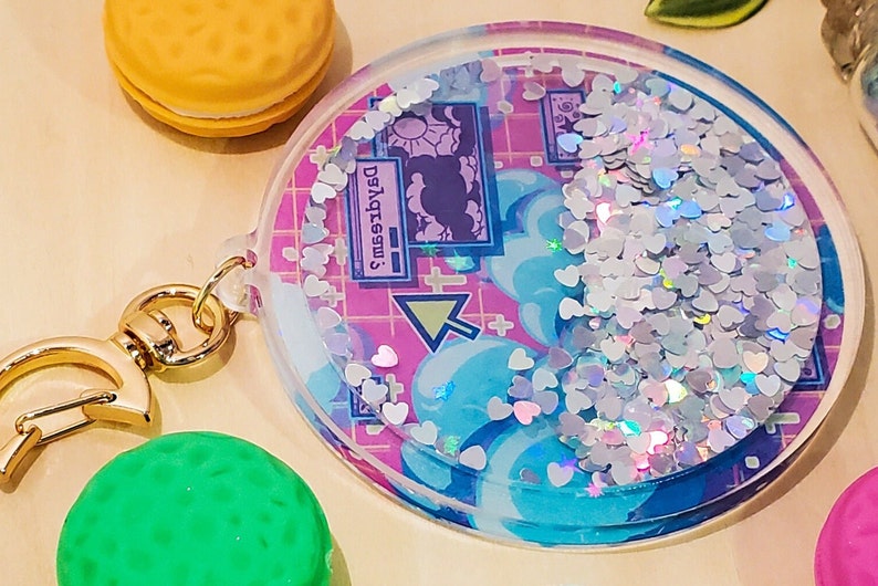 May include: A clear, round keychain with a gold clasp. The keychain features a design with blue clouds, pink grid, and heart-shaped glitter. The text "Dysgessw" is visible on the design.