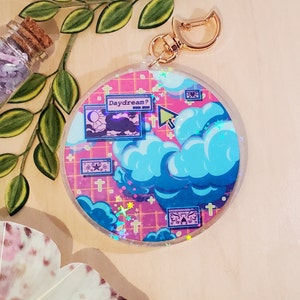 May include: A round, clear acrylic keychain with a gold clasp. The design features a pink grid background with blue clouds, stars, and the text "Daydream?". The keychain has a dreamy, whimsical aesthetic.