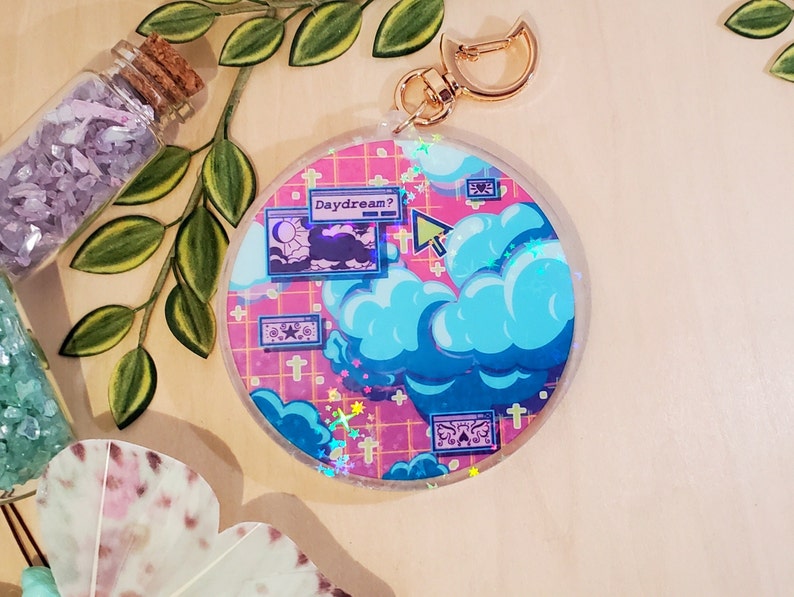 May include: A round, clear acrylic keychain with a gold clasp. The design features a pink grid background with blue clouds, stars, and the text "Daydream?". The keychain has a dreamy, whimsical aesthetic.