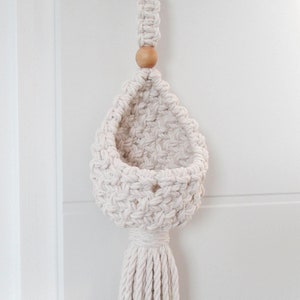 May include: A white macrame hanging planter with a wooden bead and a long tassel. The planter is made of cotton cord and has a woven design.
