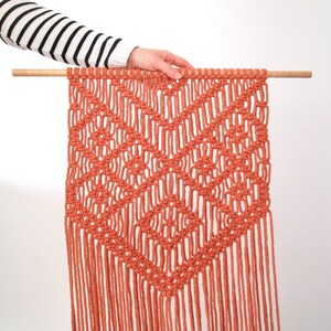 K&ouml;nnte beinhalten: Ein Wandbehang aus Makramee mit einem geometrischen Muster in Terrakotta-Orange. Der Behang ist aus dickem Baumwollgarn gefertigt und hat oben eine Holzleiste.