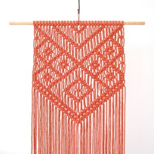 K&ouml;nnte beinhalten: Ein korallenfarbener Makramee-Wandbehang mit geometrischem Muster und Fransen. Der Behang ist aus nat&uuml;rlicher Baumwollschnur gefertigt und an einer Holzleiste befestigt.