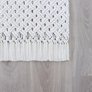 May include: A white macrame rug with a fringe border. The rug has a diamond pattern and is made of cotton cord.
