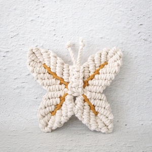 Puede incluir: Un atrapasueños de macramé blanco en forma de mariposa con detalles amarillos. La mariposa está hecha de cuerda anudada y tiene un diseño delicado e intrincado.