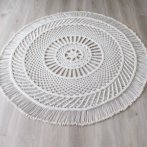Peut inclure: Un tapis rond en macram&eacute; blanc avec une bordure en franges. Le tapis a un motif circulaire au centre.