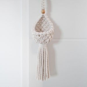 May include: A white macrame hanging planter with a wooden bead at the top and a long tassel. The planter is empty and ready to hold a small plant.